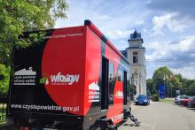 Czerwony mobilny punkt programu Czyste Powietrze zaparkowany przy ulicy, w tle budynek z wieżą zegarową.