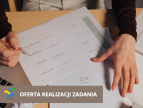 napis oferta realizacji zadania, na tle stołu na nim laptop, kartki papieru i dwie dłonie, jedma z długopisem 