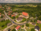 Konsulatcje społeczne planowanej budowy obwodnicy Dziekanowic Widok z lotu ptaka na malowniczą miejscowość z zabudową jednorodzinną, otoczoną zielonymi wzgórzami i polami. Na pierwszym planie widoczny kościół oraz większy budynek z czerwonym dachem, przy których krzyżują się drogi. Wokół liczne domy, drzewa i kręta szosa prowadząca przez wieś.